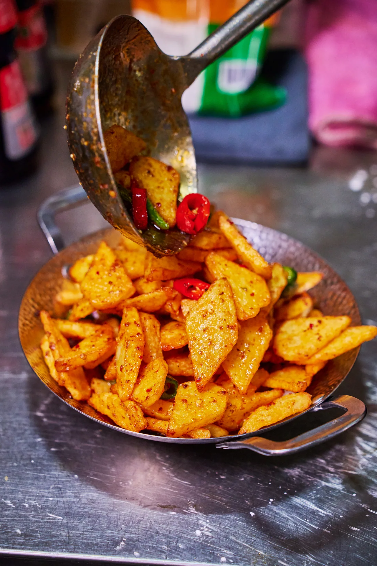 Spiced potato wedges tumbling from a ladle into a small wok with red chillies