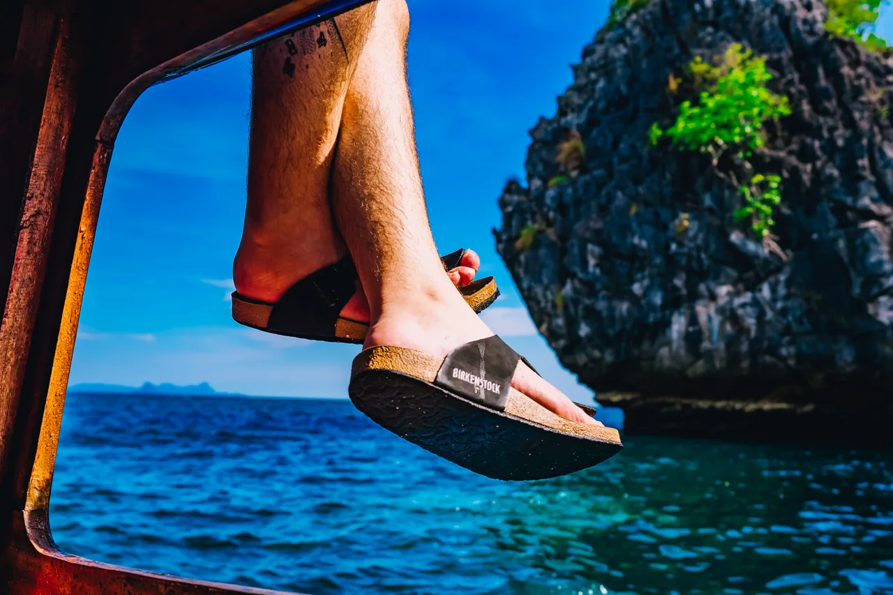 Feet hanging over the edge of a boat above a blue sea and limestone rock