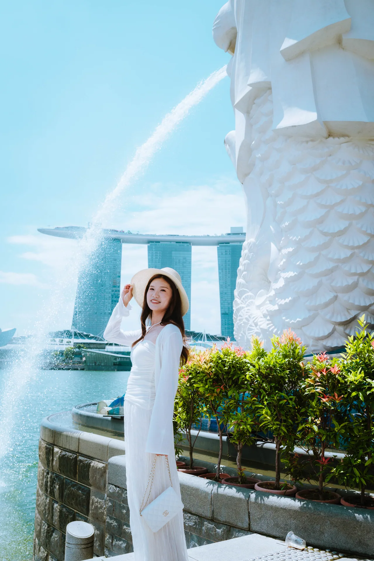 Woman in a white dress and sun hat by the Merlion, with Marina Bay Sands behind